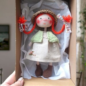 May include: A crocheted doll with red hair, a brown hat, a green cardigan, a white dress, and brown shoes. The doll is in a cardboard box with white tissue paper.