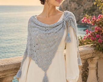 May include: A light blue triangular crochet shawl featuring a repeating scalloped pattern and decorative tassels at the corners. The shawl is draped over a plain, long-sleeved cream-coloured dress, showcasing intricate yarn work and a knotted side detail.
