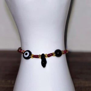 Puede incluir: Una pulsera de cordón granate que presenta una cuenta central de ojo turco en blanco y negro, un pequeño dije negro colgante y una cuenta esférica negra lisa. El diseño está acentuado con pequeñas cuentas espaciadoras doradas metálicas.
