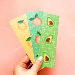 May include: Three colorful bookmarks with a grid pattern and images of lemons, peaches, and avocados. The bookmarks are made of cardstock and have a holographic finish.