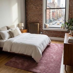 Può includere: Una camera da letto con un tappeto malva su un pavimento in legno. Il letto ha lenzuola bianche e cuscini beige. Una finestra offre una vista sulla città e una cassettiera in legno è sulla destra. Il tappeto misura circa 200 cm x 140 cm.