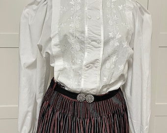 Pode incluir: Blusa branca de manga comprida com gola alta e detalhes florais bordados. A blusa tem um fecho de botões na frente e uma guarnição de renda em volta do colarinho. A blusa é combinada com uma saia de riscas pretas e vermelhas.