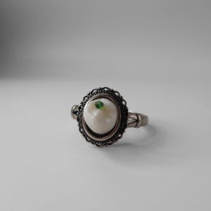 May include: A silver ring with a white tooth-shaped stone set in a filigree design. A small green gemstone is set in the center of the tooth.