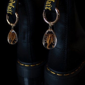 May include: A pair of black leather boots with yellow stitching. Each boot has a black strap with the text "Air Mail" in yellow letters. A silver ring with a clear teardrop-shaped pendant containing a preserved scorpion hangs from each strap.