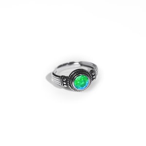 May include: A silver-toned ring featuring a central round, faceted green and blue iridescent stone. The band is crafted with a textured, oxidised metal finish, displaying intricate beaded detailing around the stone setting and grooved lines.