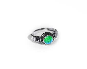 May include: A silver-toned ring featuring a central round, faceted green and blue iridescent stone. The band is crafted with a textured, oxidized metal finish, displaying intricate beaded detailing around the stone setting and grooved lines.