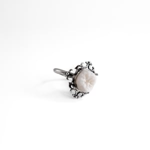 Könnte beinhalten: Ein silberfarbener Ring mit einer verzierten Metallfassung im Filigranstil, die ein einzelnes, cremeweißes, zahnförmiges Element hält. Die Ringschiene ist glatt und mit dem detaillierten, dekorativen Rahmen um das Mittelstück verbunden.