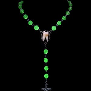 May include: A unique necklace featuring a human tooth pendant, surrounded by a silver frame, suspended from a chain of glowing green beads. A silver cross hangs at the bottom, all against a black background.
