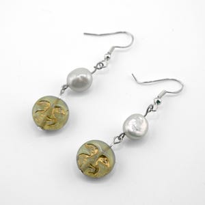 May include: A pair of drop earrings featuring silver-toned fishhook wires, each suspending a single white baroque pearl above a round, translucent pale green glass bead decorated with a gold-colored embossed celestial sun face design.