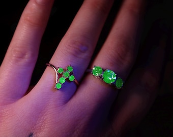 May include: Two rings with green gemstones. One ring features a cluster of green stones in a unique shape, while the other has three round green stones. The rings are set on a gold-colored band.