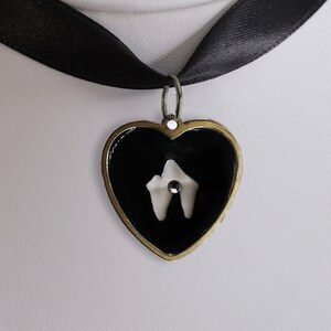 May include: A black ribbon choker necklace featuring a heart-shaped pendant with a dark metallic frame. The center of the heart is filled with black enamel and contains a small white tooth-shaped element accented with a single clear rhinestone.
