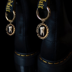 Puede incluir: Un par de botas de cuero negro con abalorios dorados atados a los cordones. Cada abalorio tiene un marco dorado con un diente blanco en su interior.