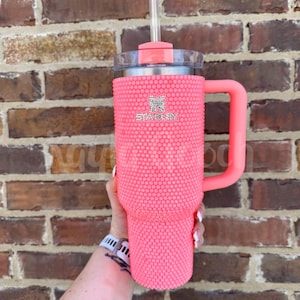 May include: A pink Stanley tumbler with a handle and straw. The tumbler is covered in small, pink, textured embellishments. The Stanley logo is visible on the front. The handle and lid are a matching pink color.