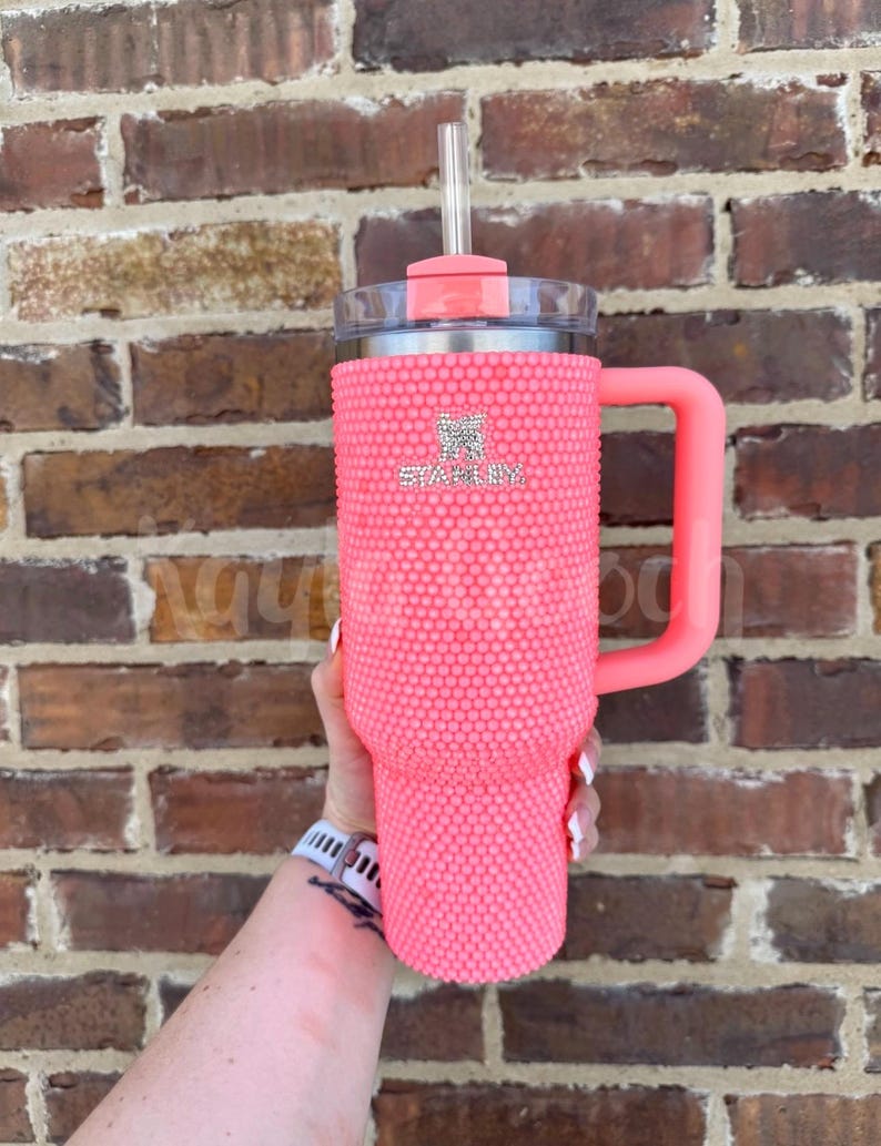 May include: A pink Stanley tumbler with a handle and straw. The tumbler is covered in small, pink, textured embellishments. The Stanley logo is visible on the front. The handle and lid are a matching pink color.