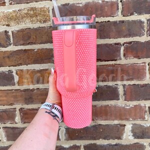 May include: A pink tumbler with a textured, studded exterior and a matching handle. The tumbler has a silver rim and a clear lid with a straw. The tumbler is held against a brick wall.