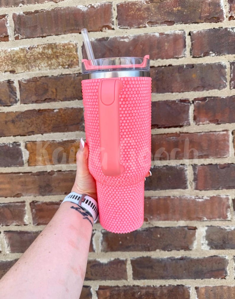 May include: A pink tumbler with a textured, studded exterior and a matching handle. The tumbler has a silver rim and a clear lid with a straw. The tumbler is held against a brick wall.