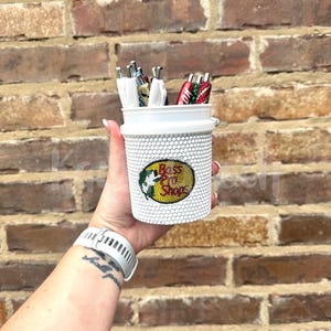 May include: A white cylindrical pen holder covered in small white beads, featuring a circular Bass Pro Shops logo patch on the front. The holder contains several pens with colorful, decorative beaded barrels in red, green, and metallic tones.