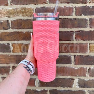 May include: A pink tumbler covered in small, round, raised embellishments. The tumbler has a silver rim and a clear lid with a straw. The background is a brick wall.