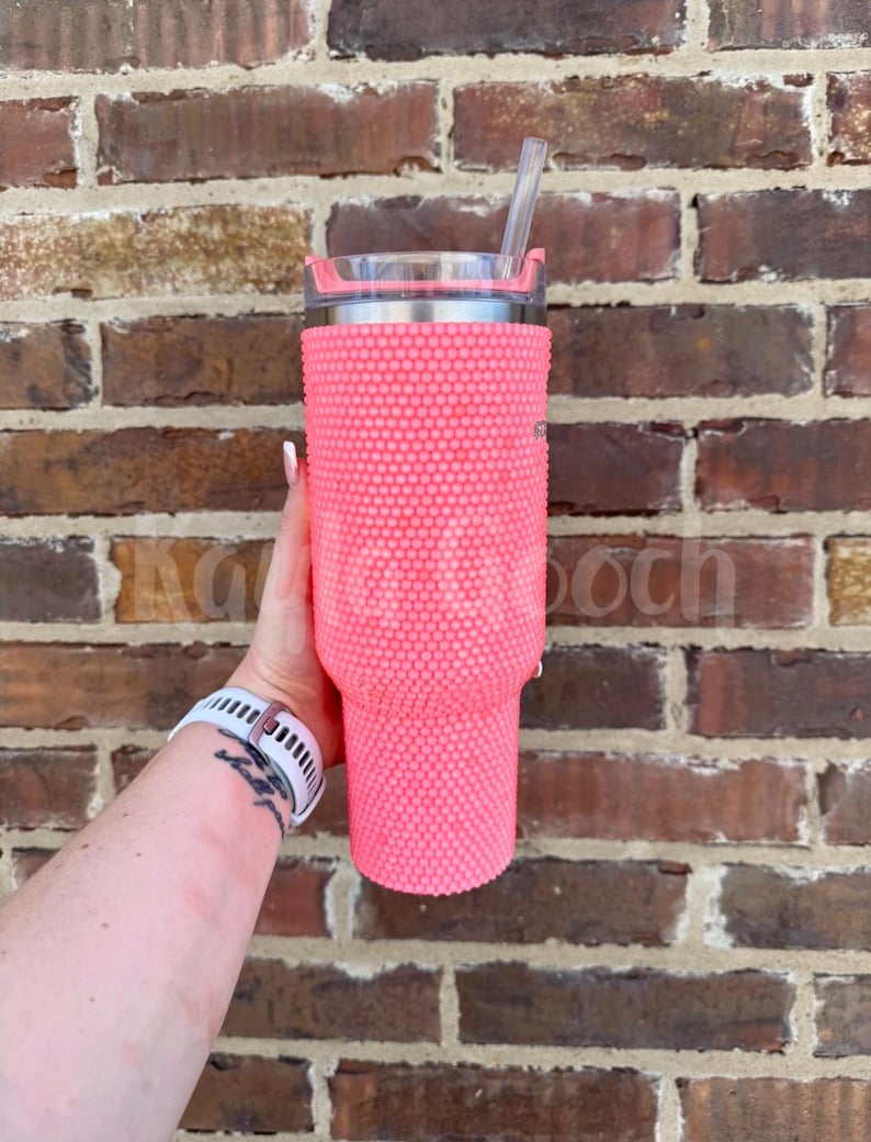 May include: A pink tumbler covered in small, round, raised embellishments. The tumbler has a silver rim and a clear lid with a straw. The background is a brick wall.