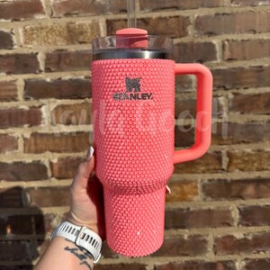 May include: A coral-colored Stanley tumbler with a textured, studded exterior. The tumbler features a handle, a clear lid, and a straw. The Stanley logo is visible on the front. The background is a brick wall.