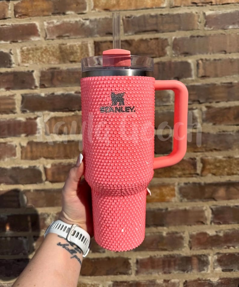 May include: A coral-colored Stanley tumbler with a textured, studded exterior. The tumbler features a handle, a clear lid, and a straw. The Stanley logo is visible on the front. The background is a brick wall.