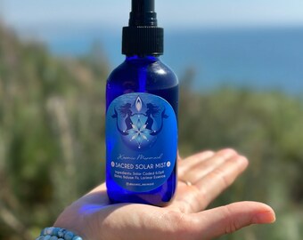 May include: A blue glass bottle with a black spray nozzle and a white label that reads "Kosmic Mermaid SACRED SOLAR MIST" with the ingredients listed below. The bottle is held in a hand with a blue and white beaded bracelet.
