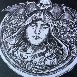 Könnte beinhalten: Eine schwarz-weiße Illustration im Tintenstil auf Stoff, die eine zentrale Figur mit langem Haar in einem Kapuzenumhang zeigt. Über dem Kopf befindet sich ein Totenkopf mit fledermausartigen Flügeln, flankiert von Blumen. Ein kreisförmiger Rahmen.
