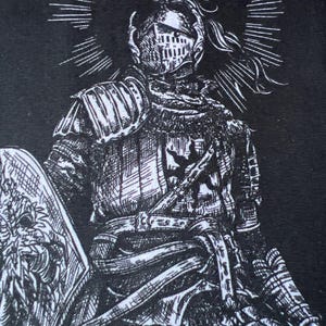 Op de afbeelding: Een zwart-witte grafische print op stof met een ridder in volledig harnas. De ridder draagt een helm met een pluim, houdt een schild met een bloemenembleem vast en heeft een zonnestraal-halo achter het hoofd, weergegeven in gedetailleerde inktlijnen.