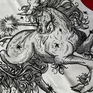 Può includere: Un quadrato di tessuto bianco con un'illustrazione dettagliata a inchiostro nero di un unicorno con criniera e coda fluenti. L'unicorno è raffigurato con una luna crescente sopra la testa, un corno luminoso e diverse stelle a quattro punte.