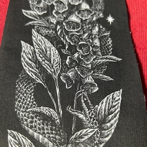 Peut inclure: Un écusson en tissu noir présentant une illustration détaillée à l'encre blanche d'un serpent enroulé entrelacé avec une tige de digitale. Le motif comprend des écailles texturées sur le serpent, des feuilles pointues et une petite étoile.