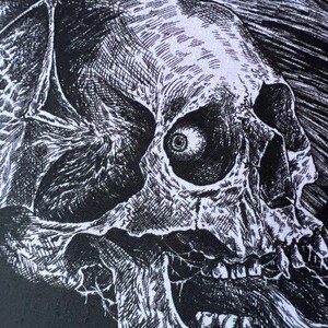 May include: A black and white graphic print on fabric featuring a detailed ink-style illustration of a human skull with a single visible eye in the socket and bat-like wings extending from the sides, set against a dark, textured background.