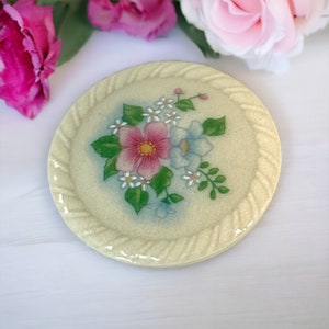 May include: A round, cream-colored ceramic coaster with a raised rope-like border. The center features a floral design with pink, blue, and white flowers.