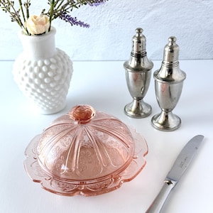 May include: A pink pressed glass butter dish with a domed lid featuring a floral pattern, two silver-toned metal salt and pepper shakers, a silver-toned butter knife, and a white hobnail vase containing a single cream flower and lavender.