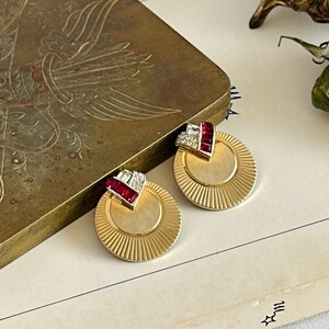 May include: A pair of gold-toned, round earrings with a sunburst design. Each earring features a V-shaped accent with red and clear gemstones. The earrings are displayed on a light-colored surface next to a vintage brass box.