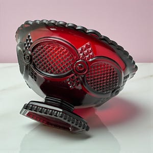 May include: A ruby red glass bowl with a pedestal base. The bowl features a diamond pattern and oval designs. The rim has a scalloped edge. The bowl is likely vintage and suitable for serving fruit or candy.