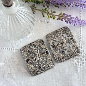 May include: Two rectangular silver-toned vintage shoe clips with intricate floral and leaf designs, encrusted with small, sparkling rhinestones. The clips are set against a white, textured background, with a glass bottle and lavender sprig in the background.