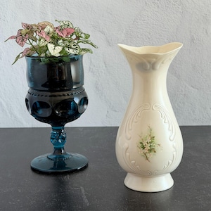 May include: A dark blue glass goblet with a textured pattern containing a small plant with pink and green leaves, positioned next to a cream-colored ceramic vase featuring a delicate floral design and embossed scrollwork on the exterior.