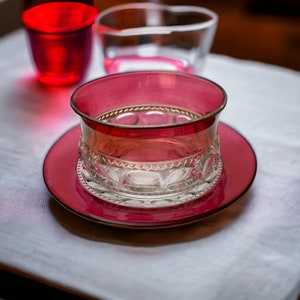 May include: A clear glass bowl with a decorative pattern sits on a red glass saucer. The bowl and saucer are both made of ruby red glass.