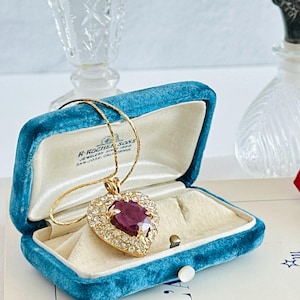 May include: A gold-toned necklace with a heart-shaped pendant featuring a large, faceted ruby surrounded by clear crystals. The pendant rests in an open, teal velvet jewelry box with a cream-colored interior. The box is labeled "R. Rocher & Sons, Jewelers, San Jose, California."