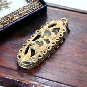 May include: An antique brass pendant with an ornate, openwork design. The pendant has a rectangular shape with a decorative border and a central design featuring swirling patterns. The pendant is likely vintage or antique, with a tarnished finish.