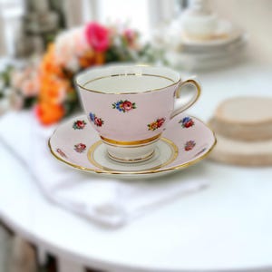 May include: A pink teacup and saucer set with floral designs and gold trim. The teacup and saucer have a delicate, vintage aesthetic, perfect for serving tea. The floral pattern includes red, blue, and yellow flowers.