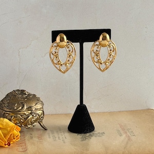 May include: A pair of gold-toned, vintage earrings. Each earring features an oval filigree design with a solid oval top. The earrings are displayed on a black stand against a neutral background. A small, ornate gold box and a yellow rose are also visible.