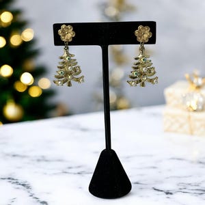 May include: Gold-toned Christmas tree earrings with pearl and green accents. The earrings are displayed on a black jewelry stand against a blurred background with Christmas lights and a gift box. The earrings have a flower at the top.