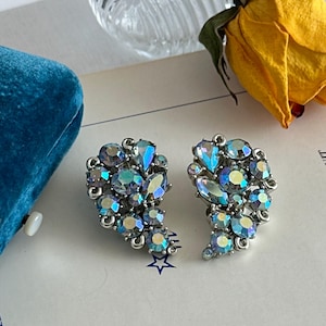 May include: A pair of vintage-style, silver-toned clip-on earrings. Each earring features an arrangement of iridescent, oval, and teardrop-shaped rhinestones in shades of blue, purple, and green. The earrings are set against a white background with a blue velvet surface.