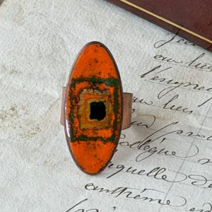 May include: An oval-shaped ring featuring a vibrant orange enamel face with concentric rectangular patterns in shades of yellow, green, and a solid black center. The band is constructed from a plain, smooth copper-toned metal material.