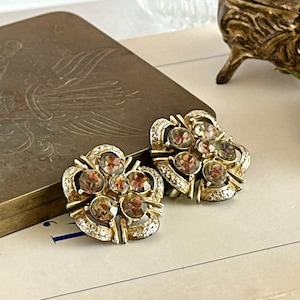 May include: A pair of vintage gold-toned clip-on earrings. Each earring features a floral design with multiple brown-toned gemstones and clear crystals. The earrings are set against a neutral background.