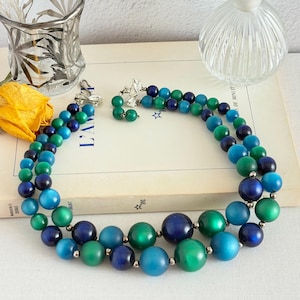 May include: A vintage two-strand beaded necklace in shades of blue and green. The necklace features large, round beads in varying sizes and hues, with a silver-tone clasp. The necklace is displayed on a book with the word "L'AM" visible.