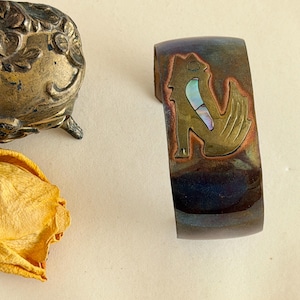 以下が含まれることがあります： 暗く虹色の仕上げが施された、幅広の金属製カフブレスレット。アワビの象嵌が施された、金色の様式化された動物のデザインが特徴です。カフはニュートラルな背景に表示されています。