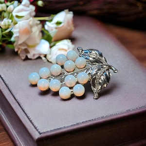 May include: A silver grape cluster brooch with white opal stones. The brooch features a detailed silver leaf design.
