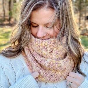 May include: A woman wearing a white cable knit sweater with a pink and yellow crocheted infinity scarf.